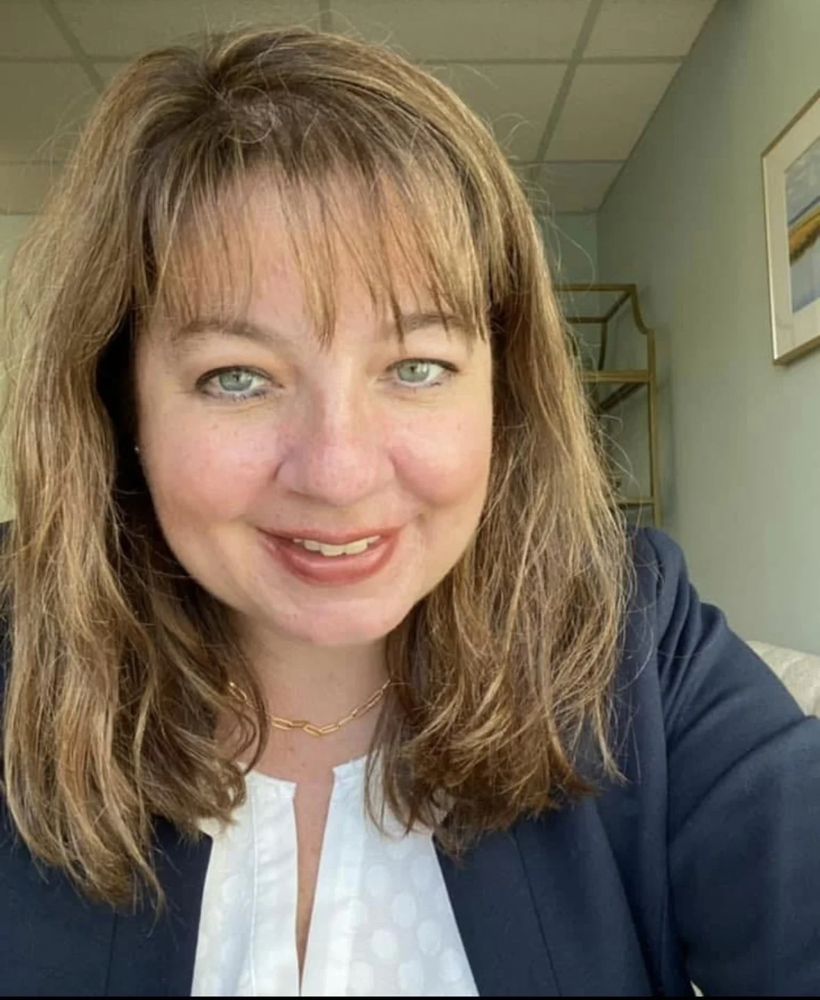 A woman with shoulder-length light brown hair, blue eyes, and light skin smiling at the camera, wearing a navy blazer over a white blouse, with a gold necklace, in an indoor setting with light-colored walls and a framed picture in the background.