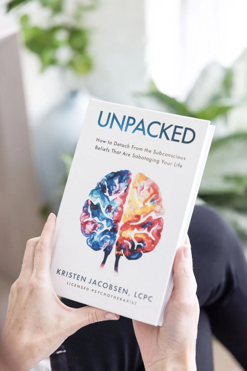 A person holding a book titled "UNPACKED" by Kristen Jacobsen, LCPC, with a colorful illustration of a human brain on the cover, in a bright room with green plants in the background.
