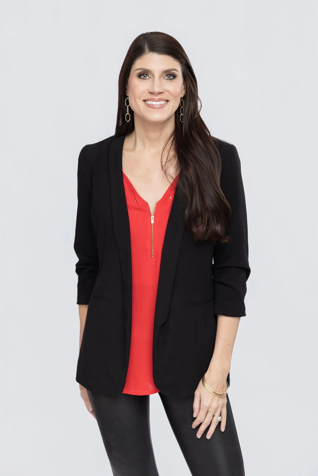 A woman with long dark hair, wearing a black blazer over a red top, and black pants, standing against a plain white background and smiling.