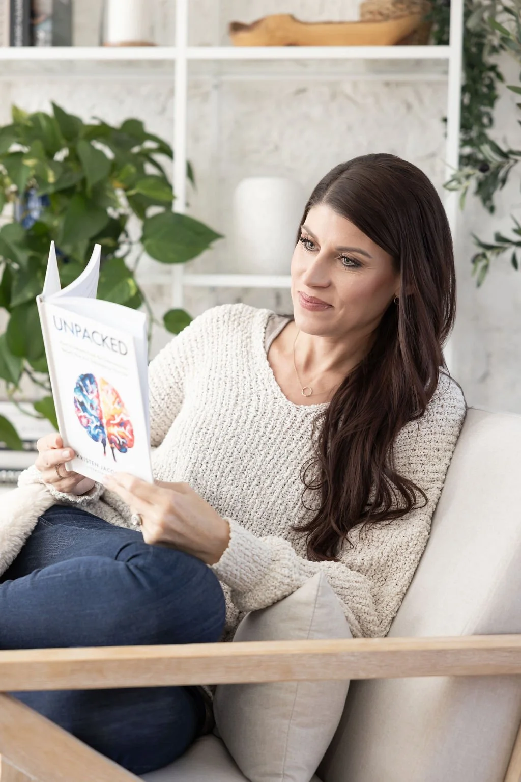 Woman with long brown hair sitting on a light-colored sofa, reading a book titled 'Unpacked' featuring a brain illustration on the cover, in a bright room with plants and white shelving in the background.