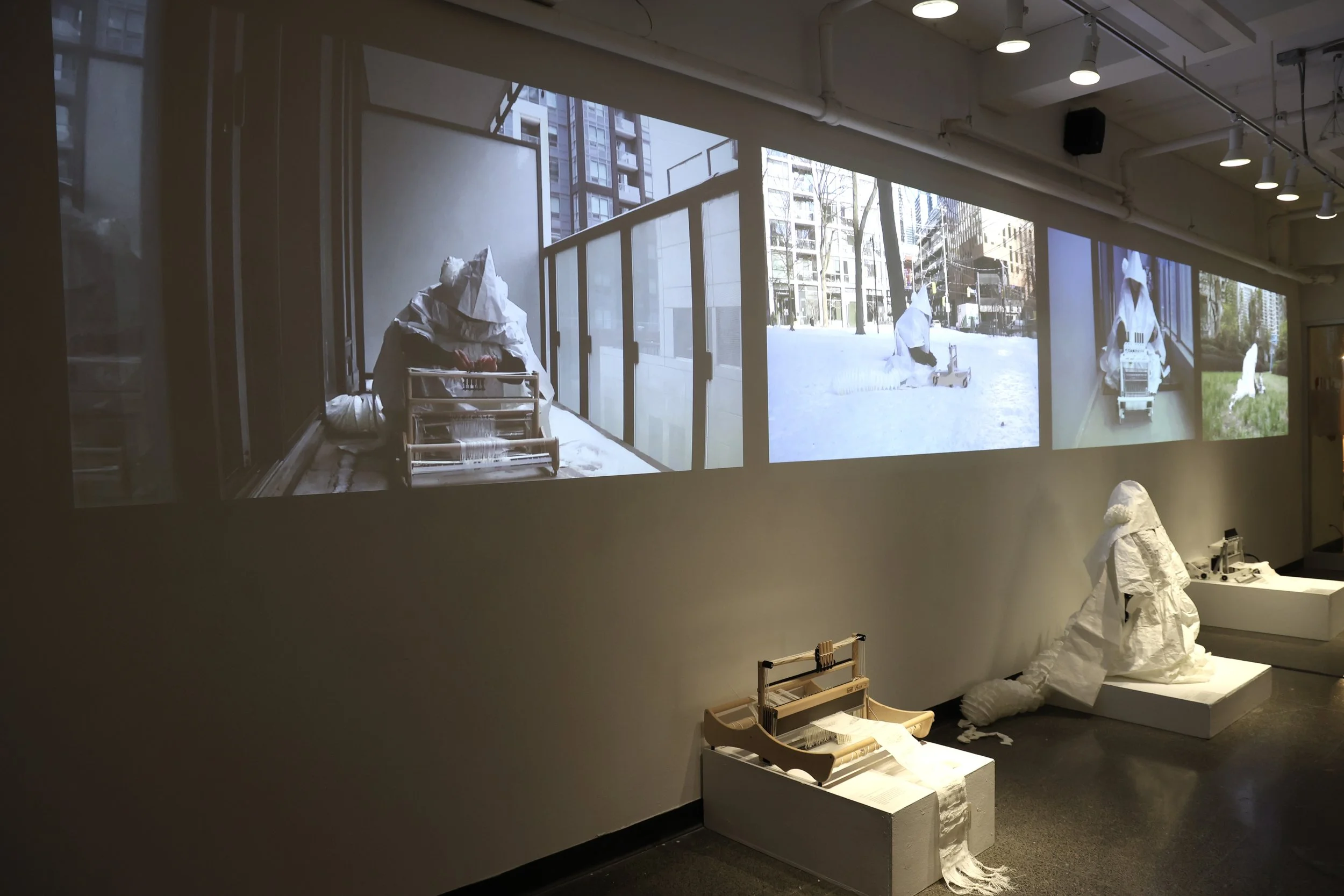 Installation view of the exhibition hallway, showing four projected performance videos displayed simultaneously alongside the loom, woven cloth, and costume. Photograph by Aritra Das, 2026.