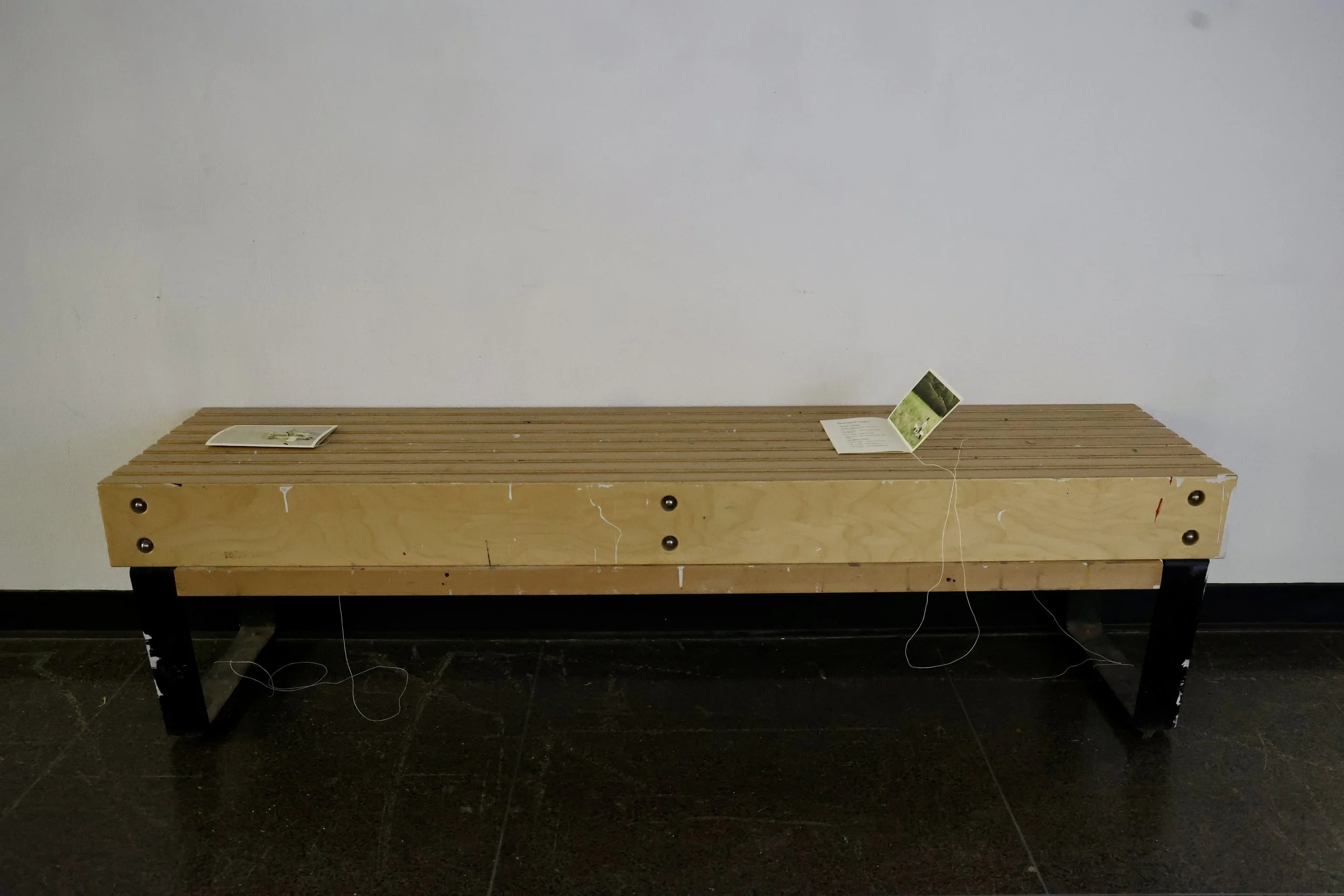 Installation view of the hallway bench with printed booklets tied in place using washi thread, providing viewers with contextual material to read while watching the videos. Photograph by Aritra Das, 2026.