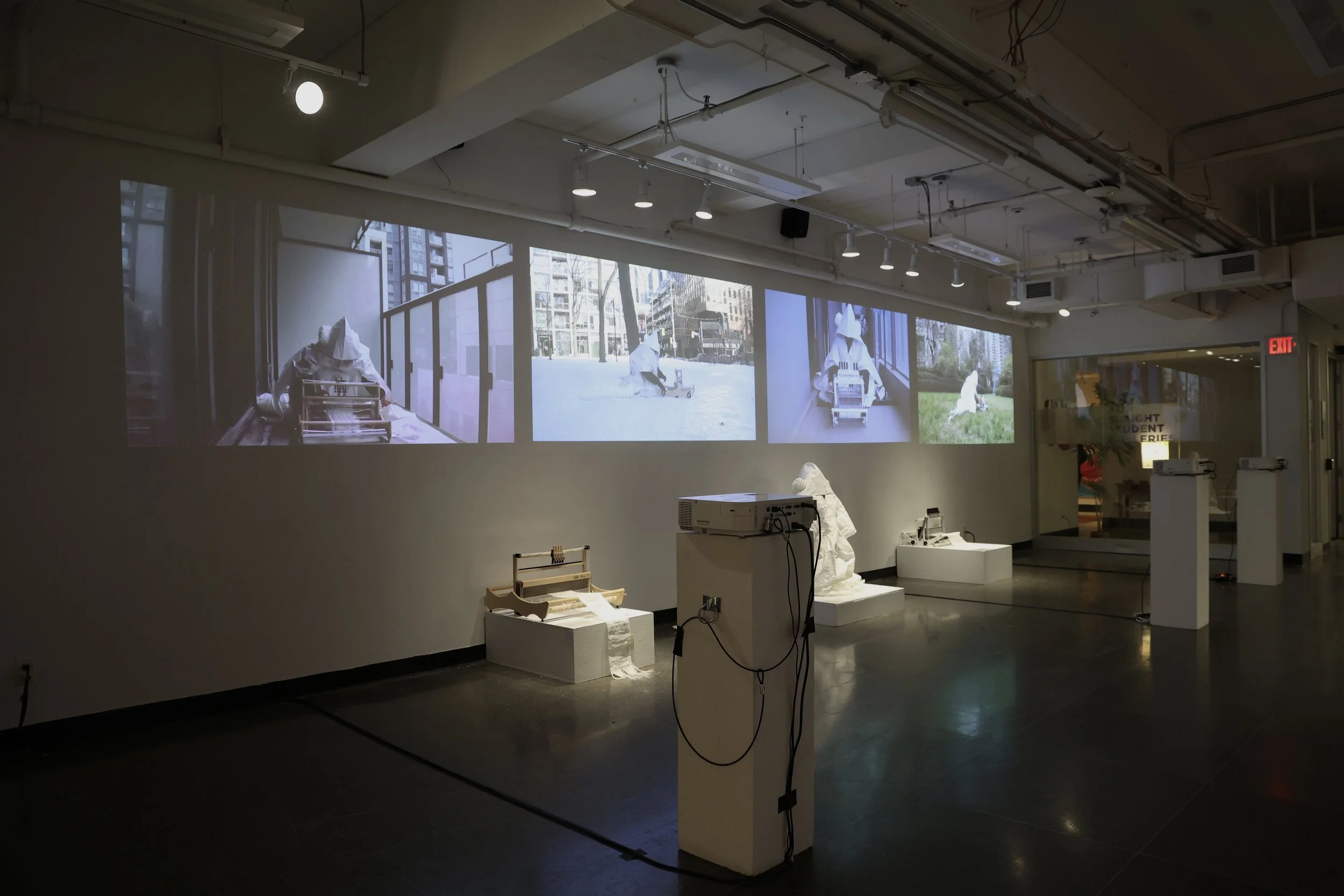 Installation view of the exhibition hallway, showing four projected performance videos displayed simultaneously alongside the loom, woven cloth, and costume. Photograph by Aritra Das, 2026.
