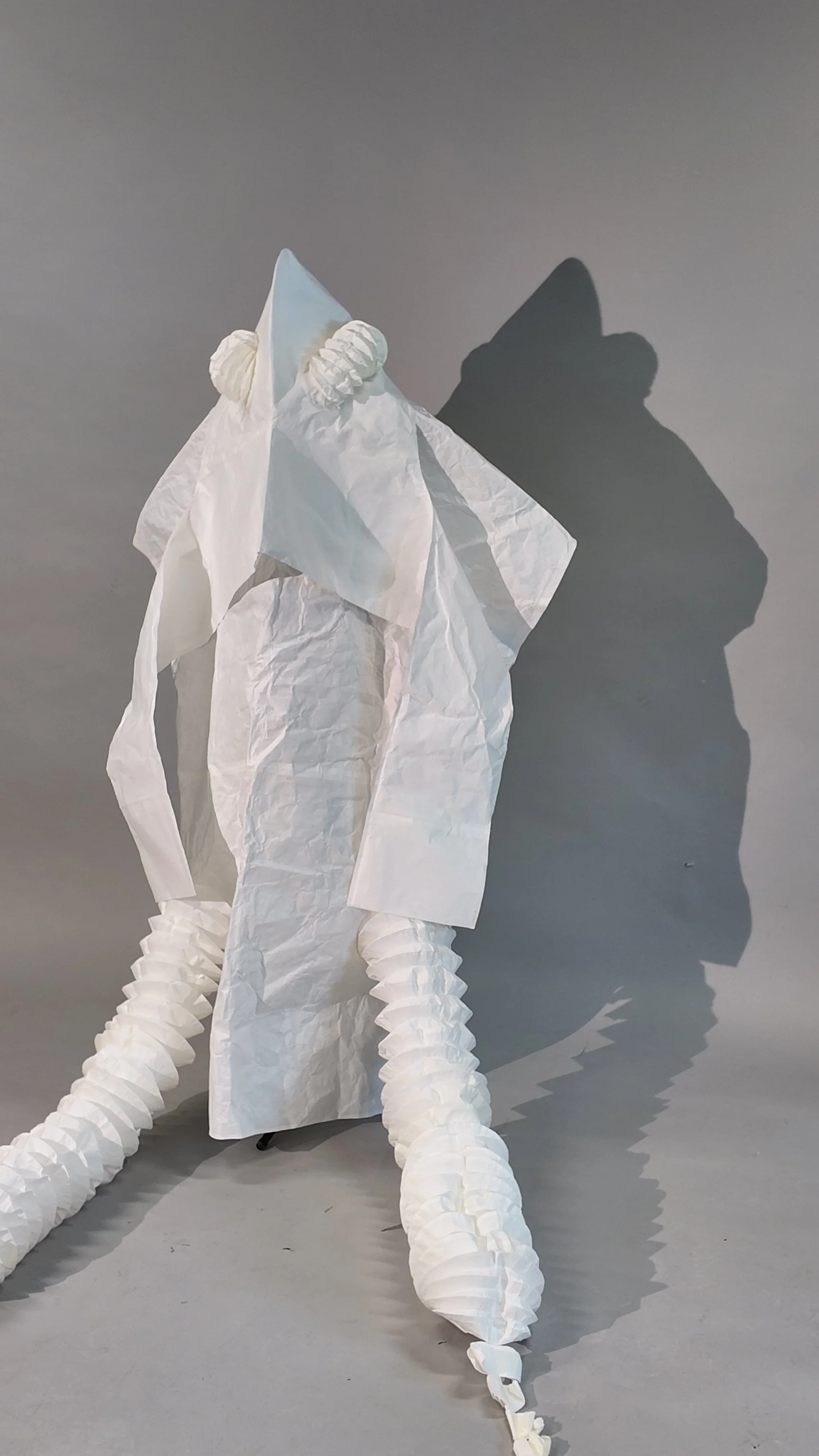 Back view of the final white washi paper squid-yokai costume, showing the two extended tentacles attached opposite each other along the back and the long trailing hood extensions. The cast shadow reinforces the squid-like silhouette. Photograph by Yu