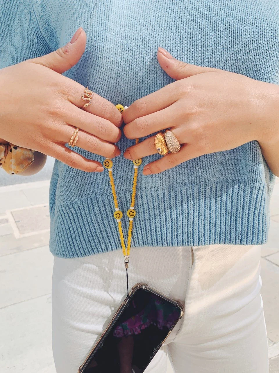 Person wearing multiple rings, holding a yellow beaded phone charm with smiley face beads, The person is dressed in a blue sweater and white pants.