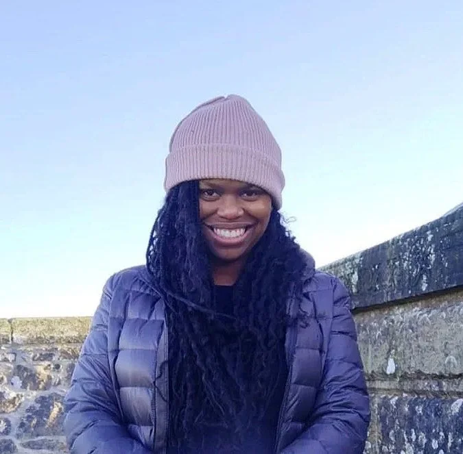 A woman smiling outdoors in front of a stone wall, wearing a pink beanie, a black shirt, and a purple puffer jacket.