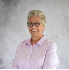 Smiling person with short gray hair and glasses wearing a lavender polka dot blouse against a gray background.