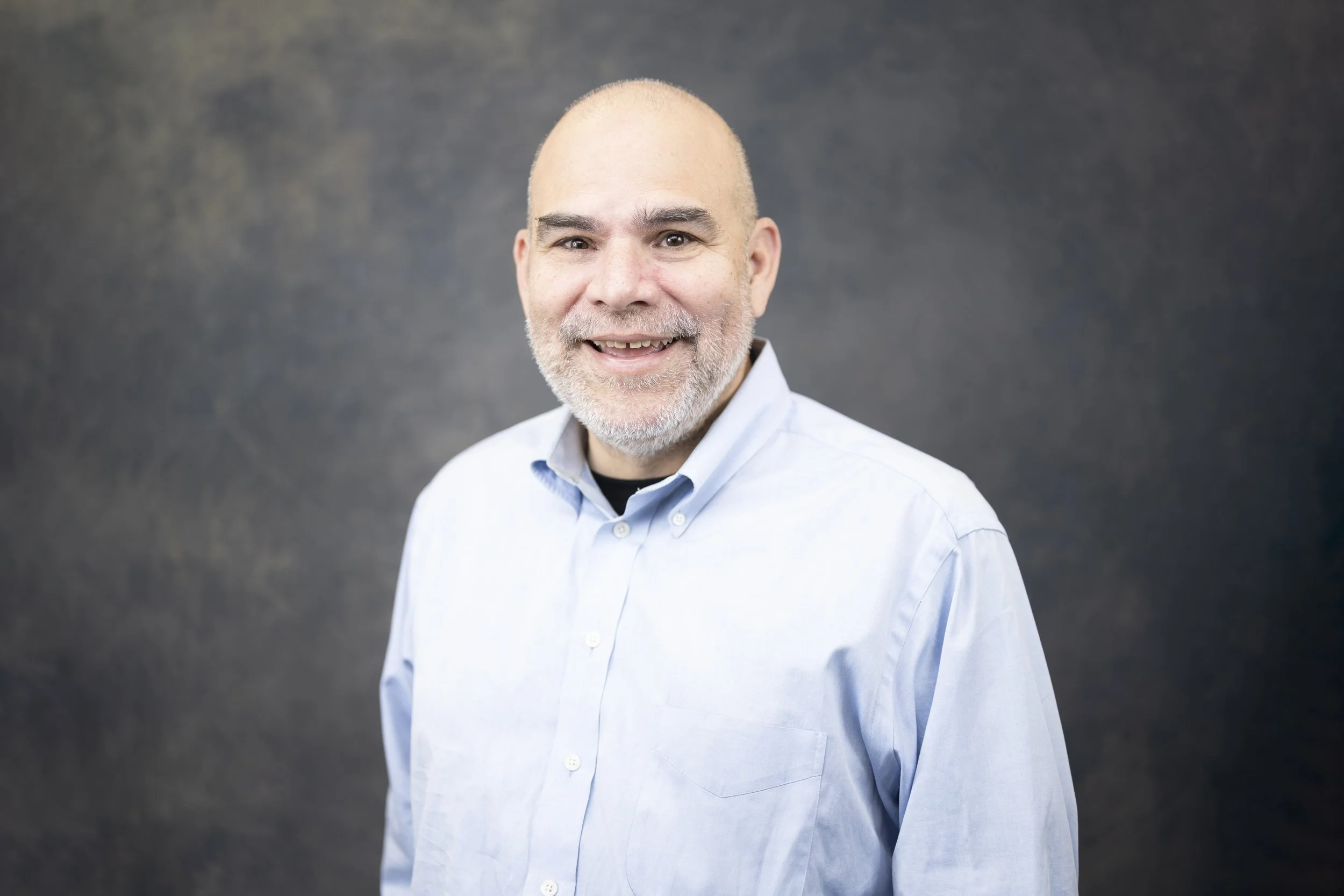 A middle-aged man with a gray beard, wearing a light blue button-up shirt, smiling against a dark gray background.