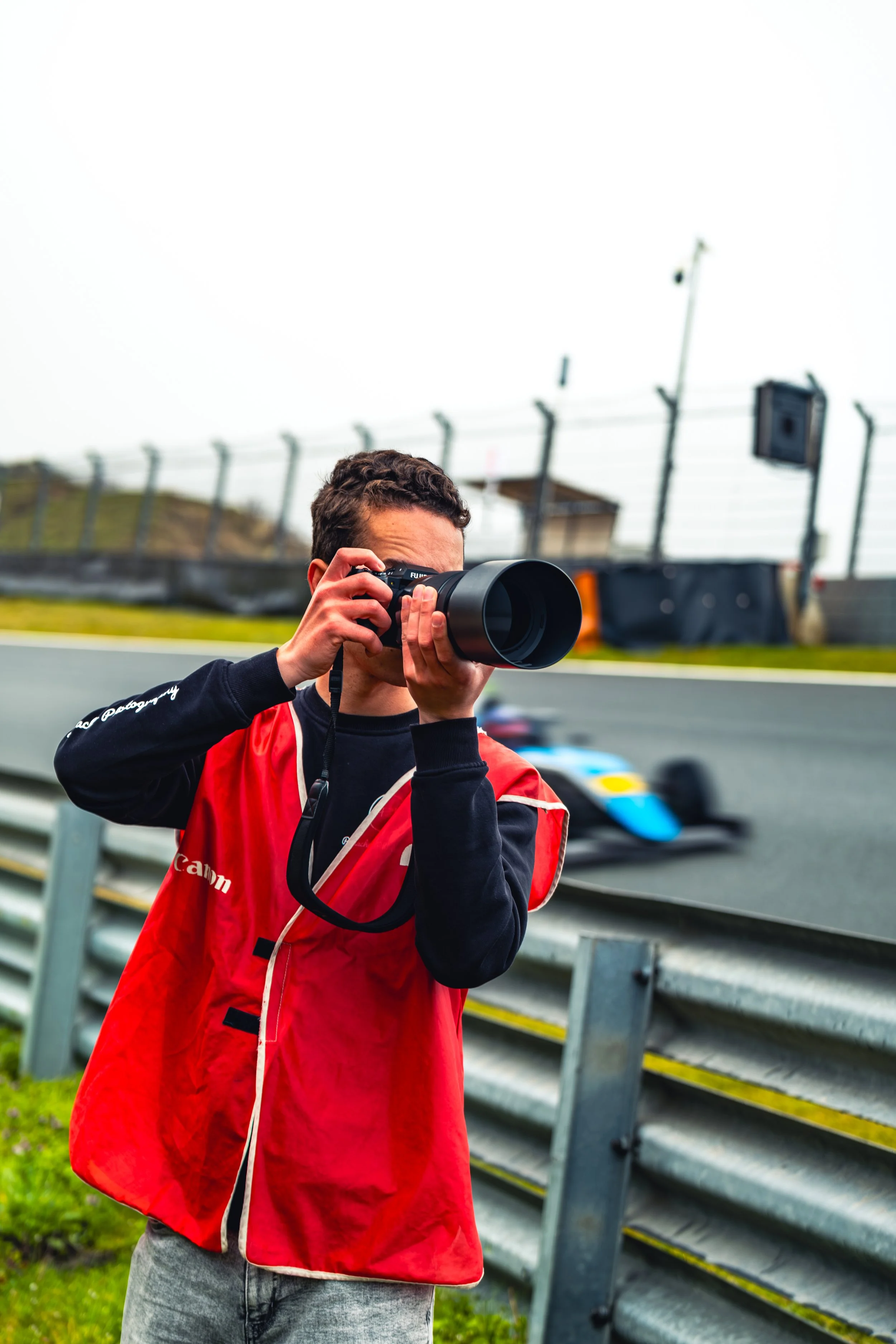 A person holding a camera at a race track, wearing a red jacket and gray sweatpants, with parade cars passing by in the background.