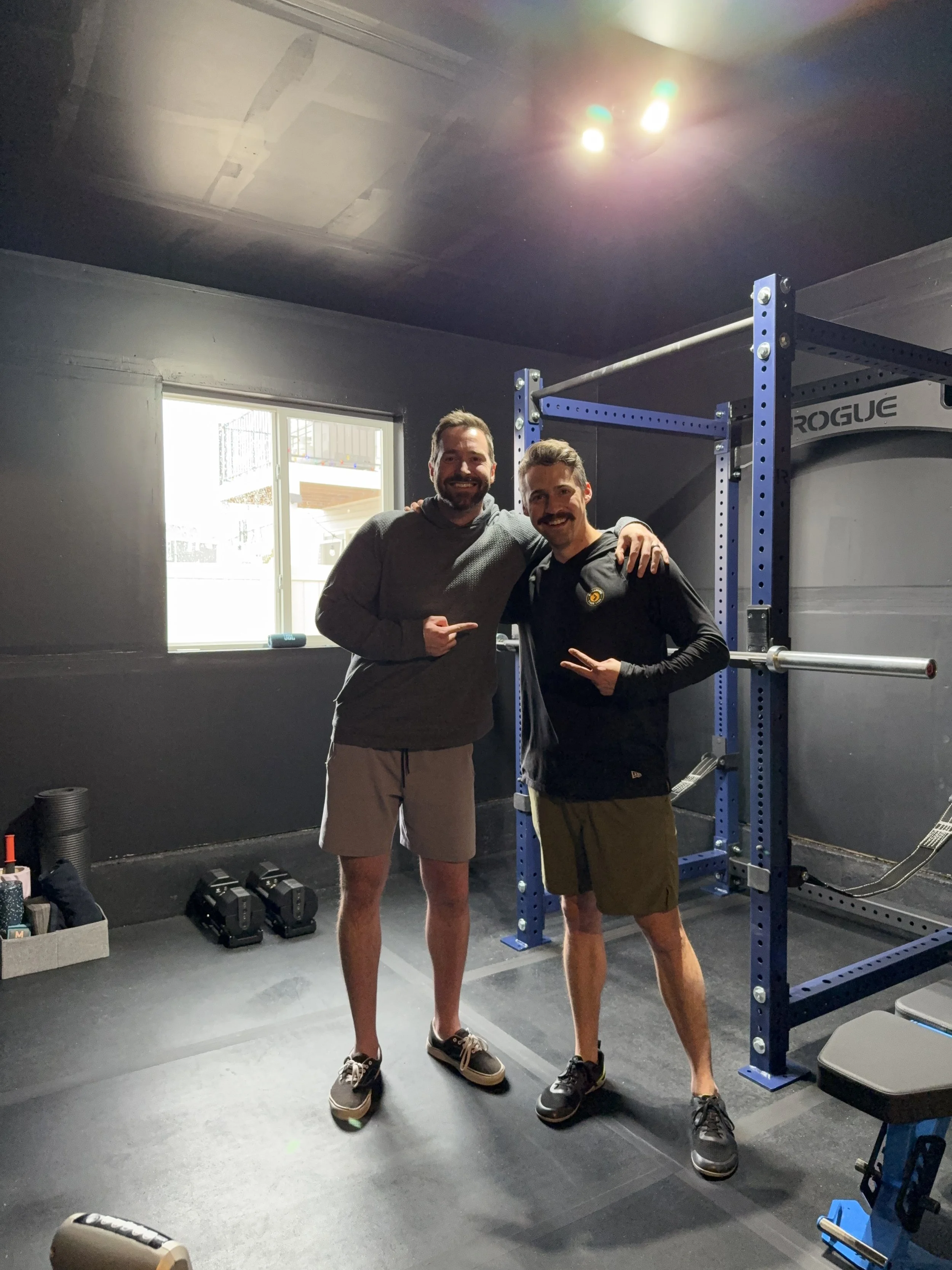 Two men smiling and posing for a photo in a home gym, standing next to a blue squat rack. One man wears a gray hoodie and shorts, the other a black jacket and shorts. The gym has black walls, a window, and various fitness equipment on the floor.