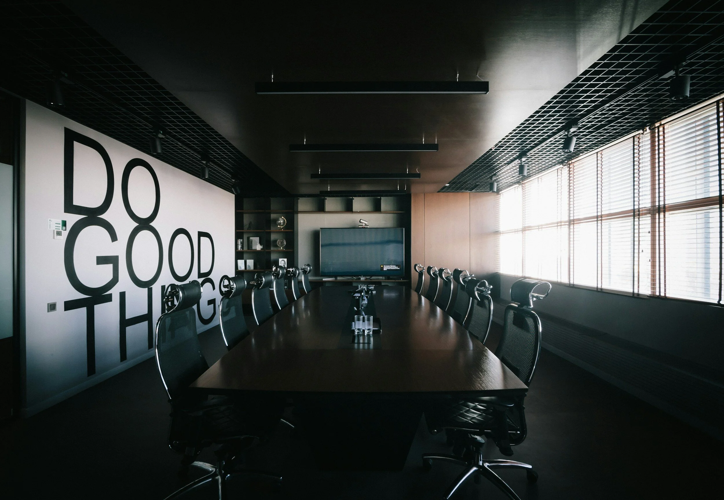 Empty boardroom with a large wooden table, black swivel chairs, a flat screen TV, and a wall with the words 'DO GOOD TH' in large black letters, with sunlight coming through window blinds on the right.