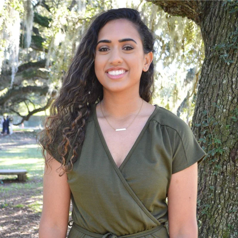 A young woman with long curly dark hair smiling outdoors near a tree.