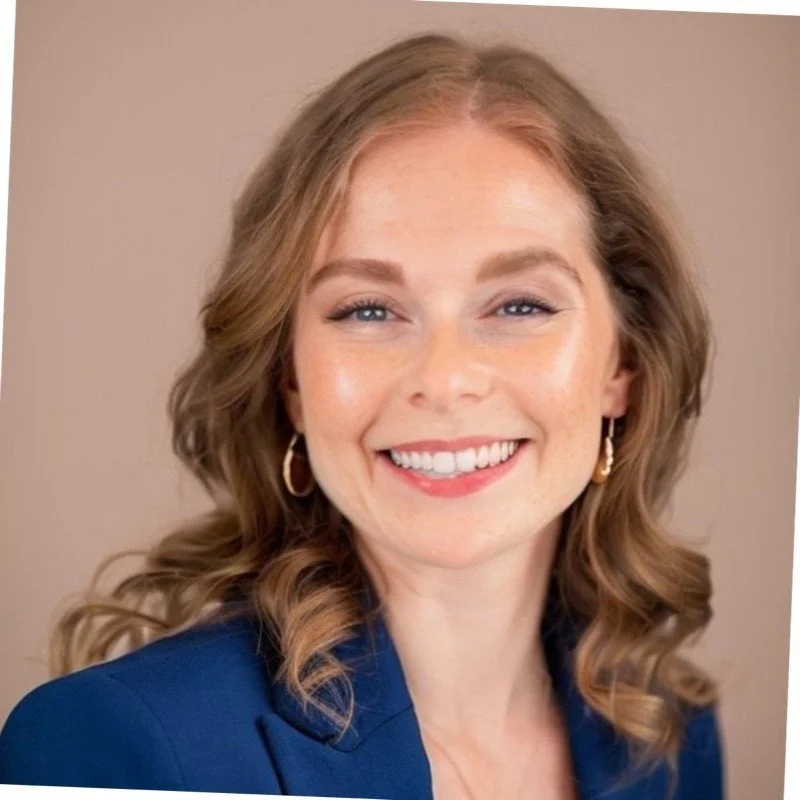 A woman with wavy, shoulder-length red hair, blue eyes, wearing earrings and a blue blazer, smiling at the camera.