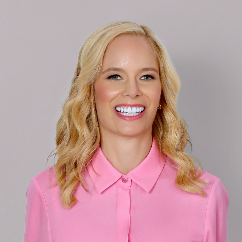 A woman with blonde wavy hair wearing a pink collared shirt, smiling against a plain light gray background.