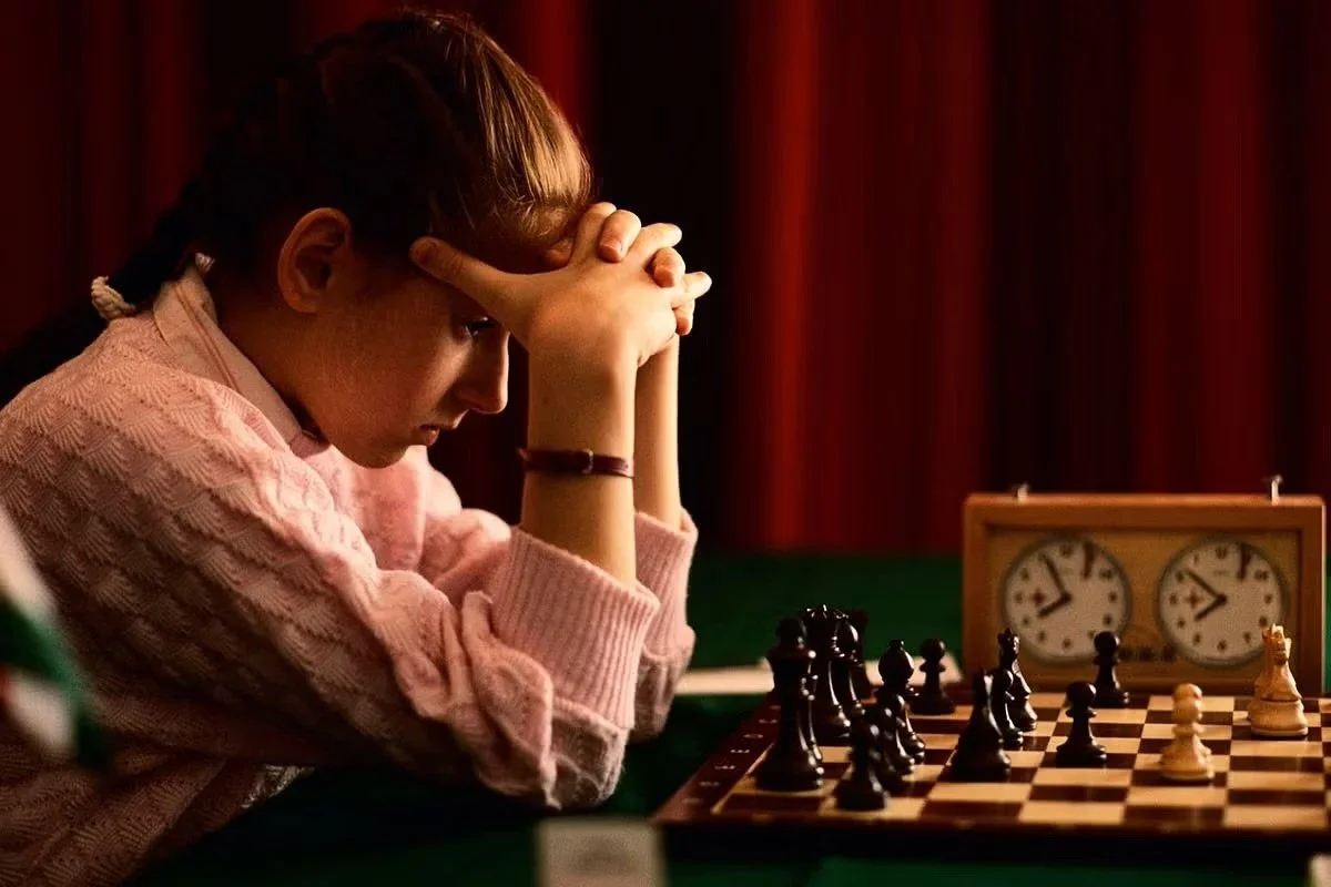Young woman playing chess, sitting at a table with chess pieces, a chess clock, and a green tablecloth, with a red curtain in the background.