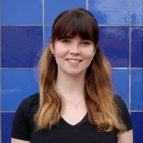 A young woman with long, wavy hair and bangs, wearing a black shirt, standing in front of a blue tiled wall and smiling at the camera.