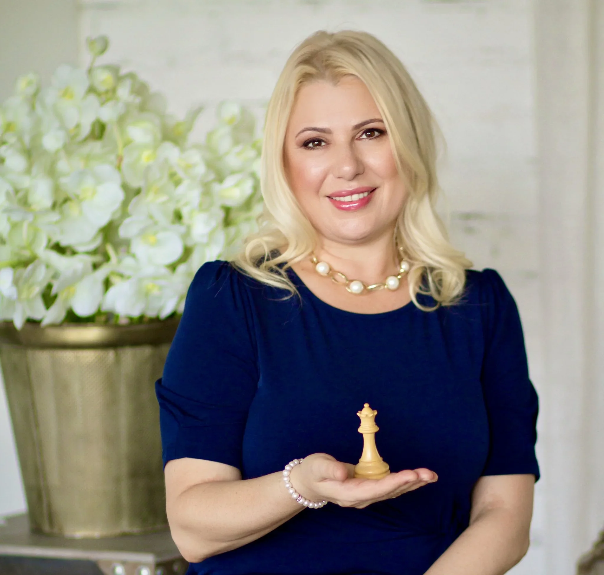 A woman with blonde hair, wearing a dark blue dress and pearl jewelry, smiling and holding a small chess queen piece in her hand, with white flowers in a vase in the background.