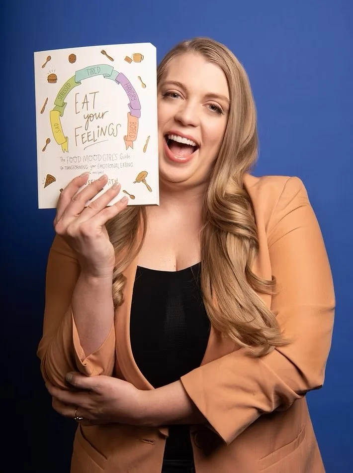 A woman with long blonde hair smiling and holding a booklet titled 'Eat your feelings' with a colorful circular diagram and illustrations of food around her, standing against a blue background.