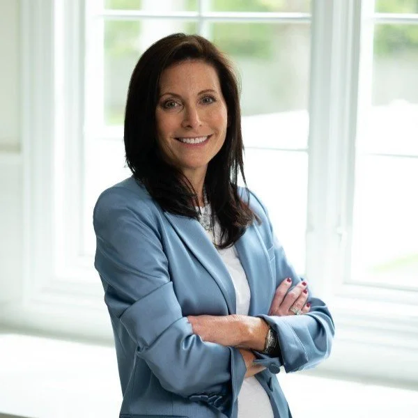 A woman with dark brown hair smiling, wearing a light blue blazer and white top, standing in front of large windows with a view of greenery outside.