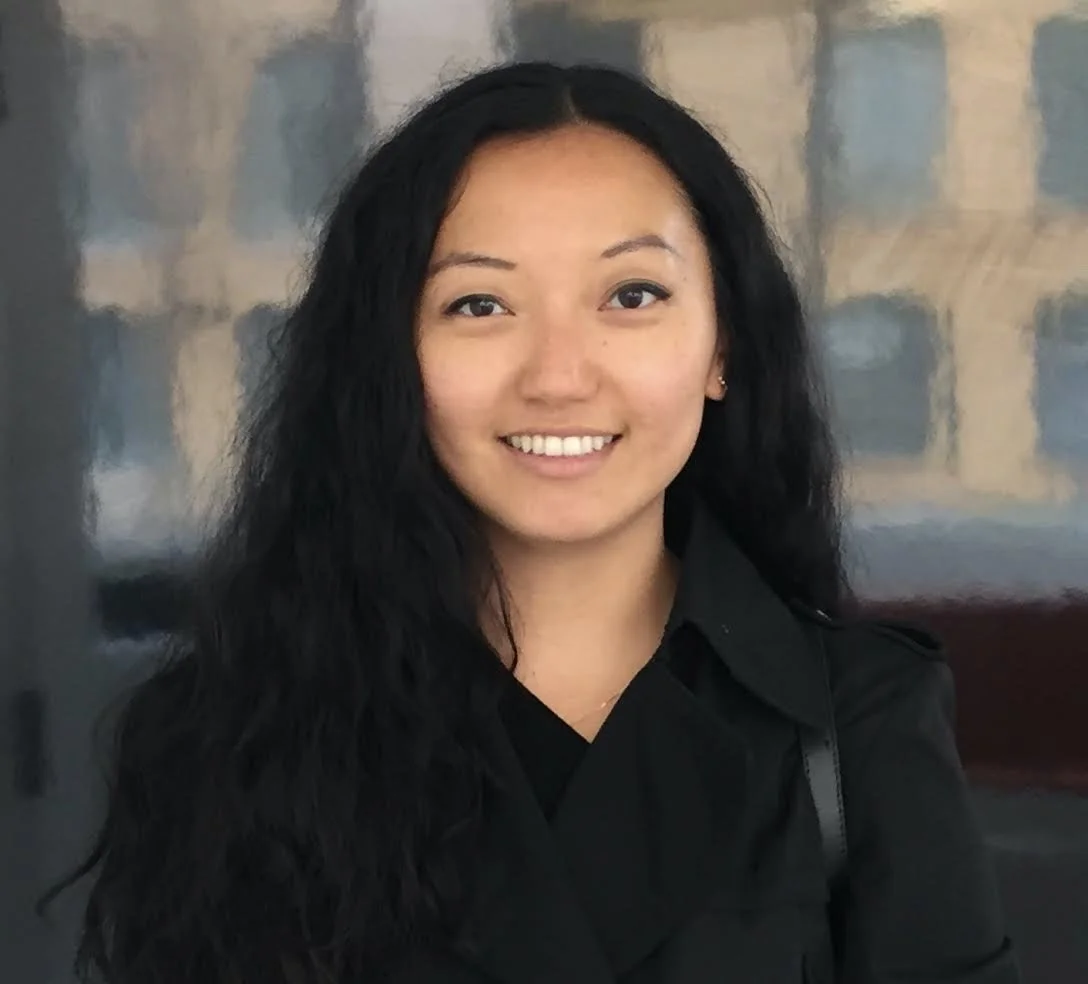 A young woman with long, dark, wavy hair smiling, wearing a black shirt, standing indoors with a blurred abstract painting or wall art in the background.