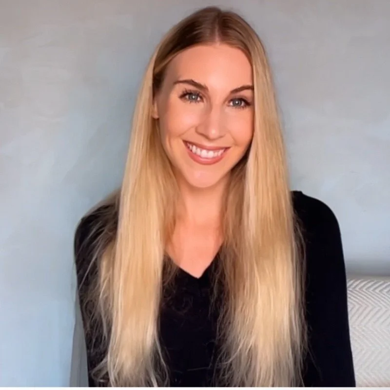 A young woman with long blonde hair, blue eyes, and a broad smile, wearing a black top, sitting against a light blue wall.