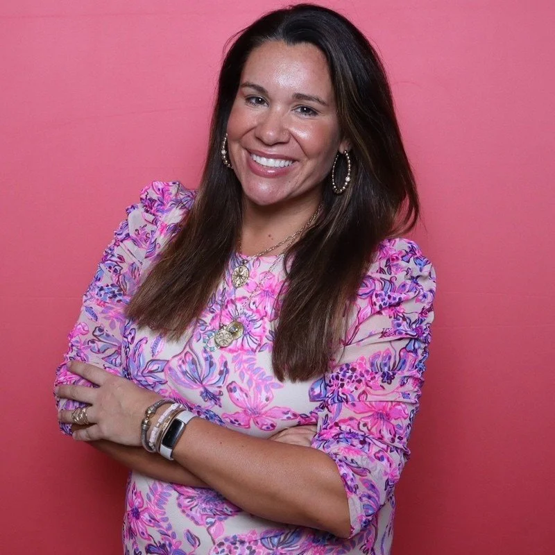A woman with long brown hair, smiling, wearing a pink and purple floral top, gold earrings, layered necklaces, and bracelets, standing against a pink background.