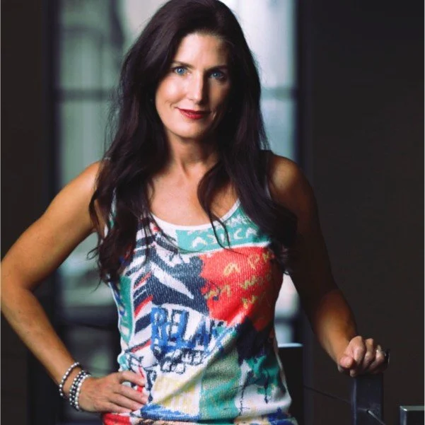 A woman with long dark hair and blue eyes wearing a colorful sleeveless top, standing indoors with a confident smile.