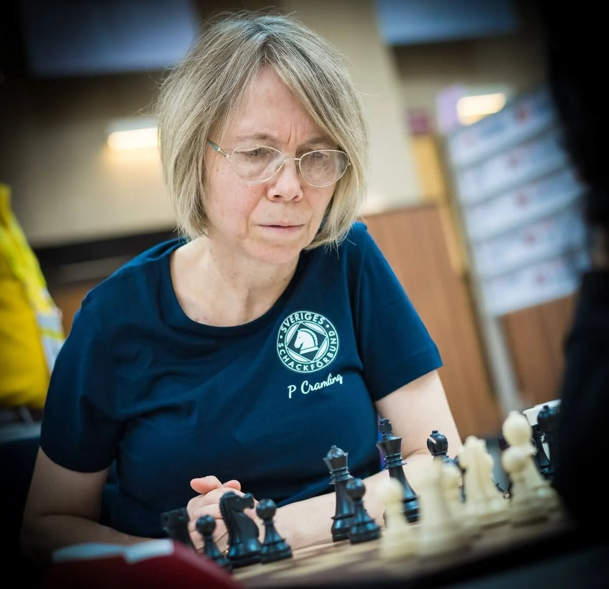 A middle-aged woman with glasses playing chess indoors, wearing a navy blue shirt with a logo and the name P Cramling.