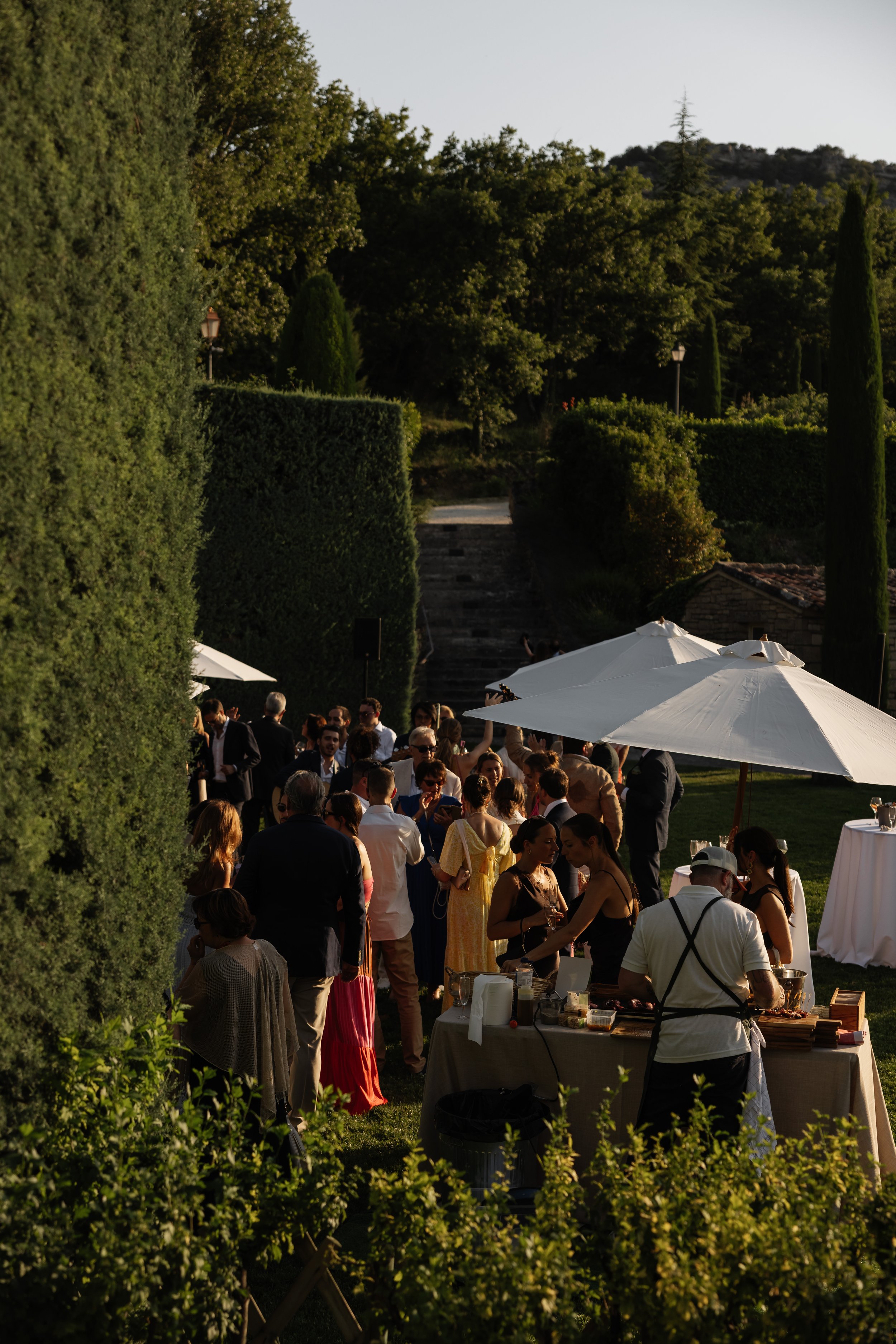 Outdoor gathering with guests under umbrellas, surrounded by lush green trees and bushes, with steps leading up a hill in the background. Event shot in the South of France