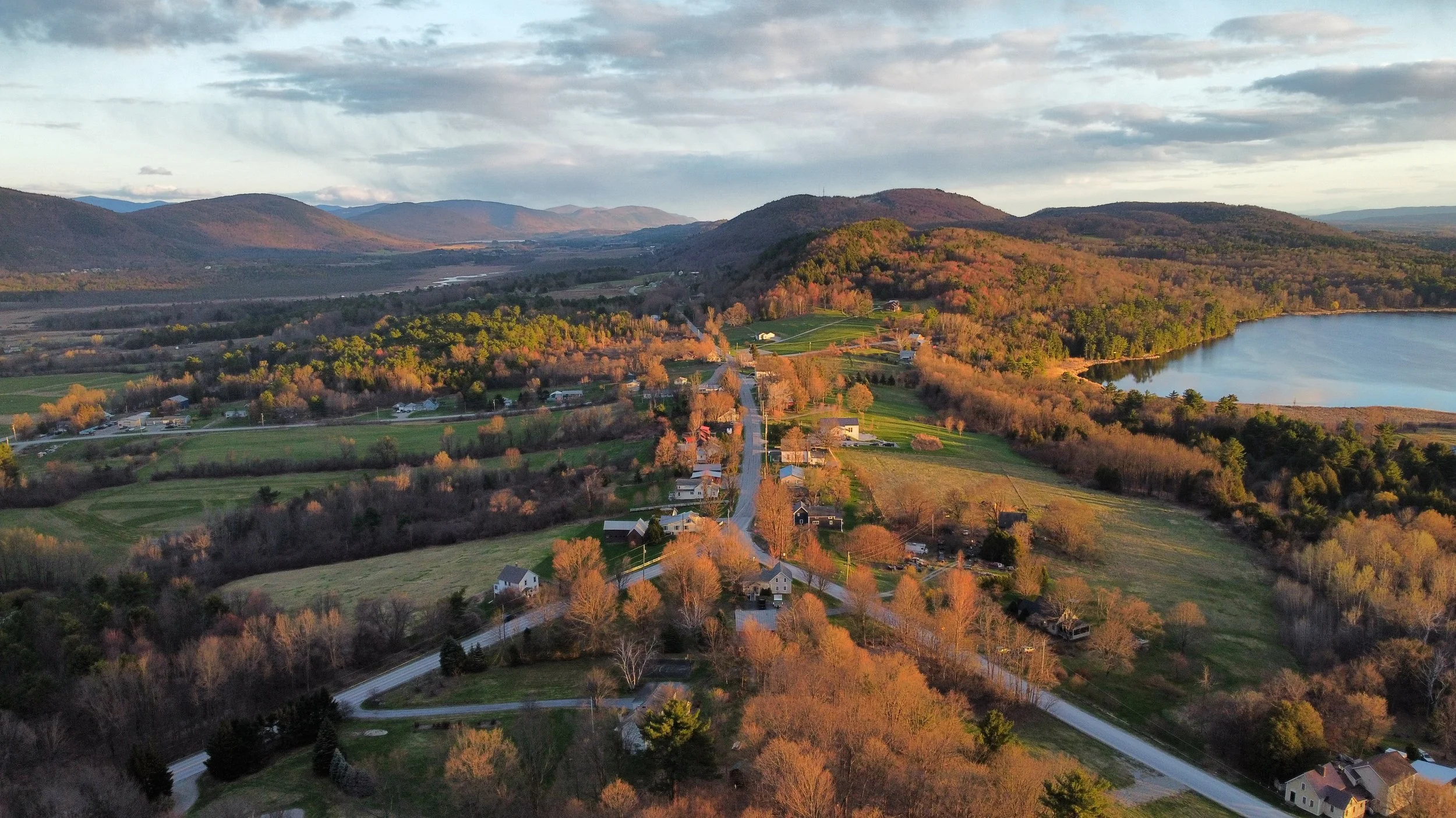 Aerial view of a Vermont valley at autumn peak, with rolling hills, two roads converging in the foreground, open farmland, and a lake in the distance beneath a dramatic cloudy sky.