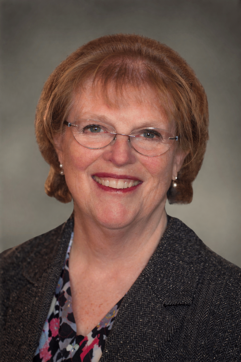 Professional headshot of Linda, RN, a woman with short auburn hair and glasses, smiling warmly in a blazer against a neutral gray background.