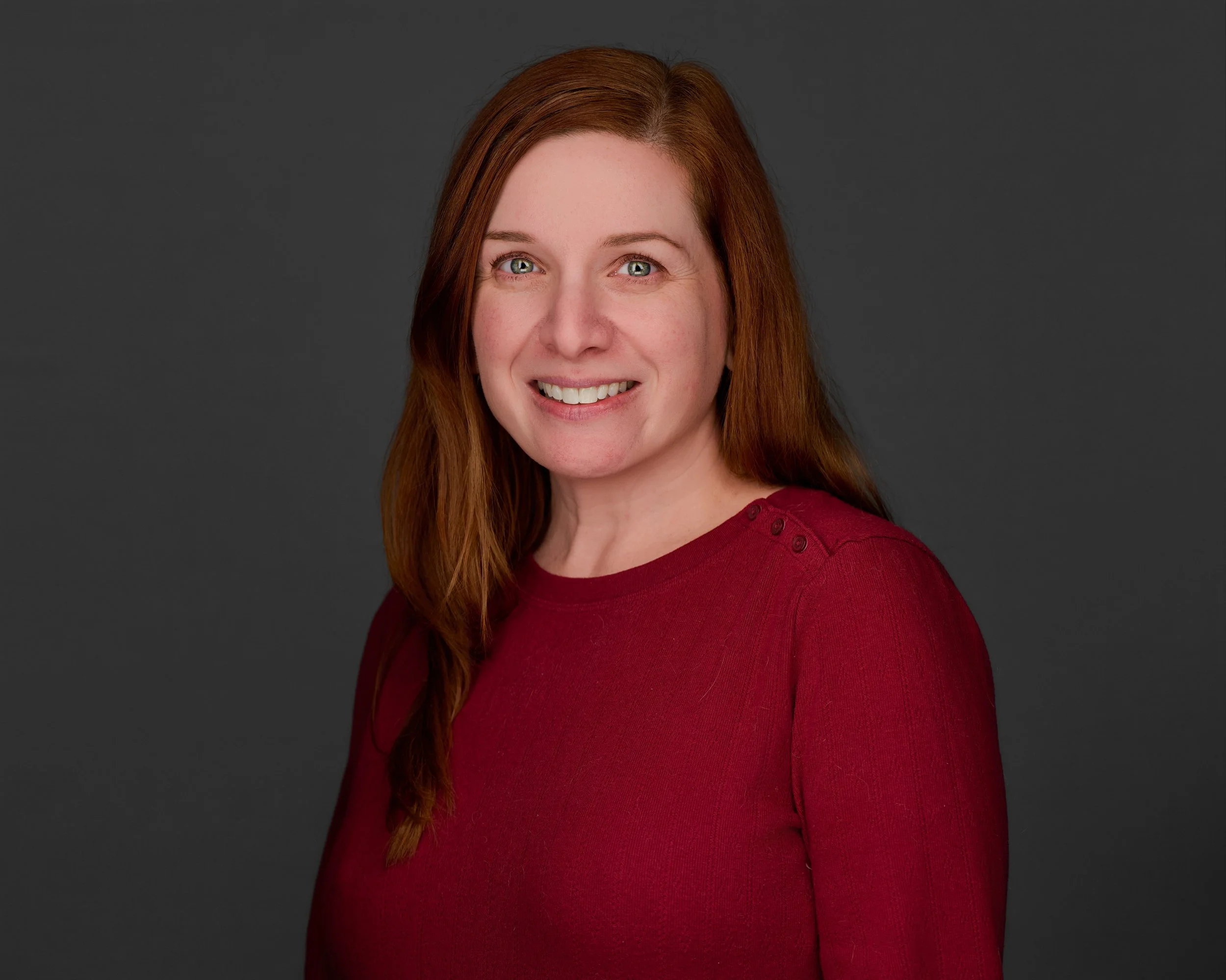 Professional headshot of Melissa Jarmel, a woman with long auburn hair and green eyes, smiling warmly in a red top against a neutral gray background.