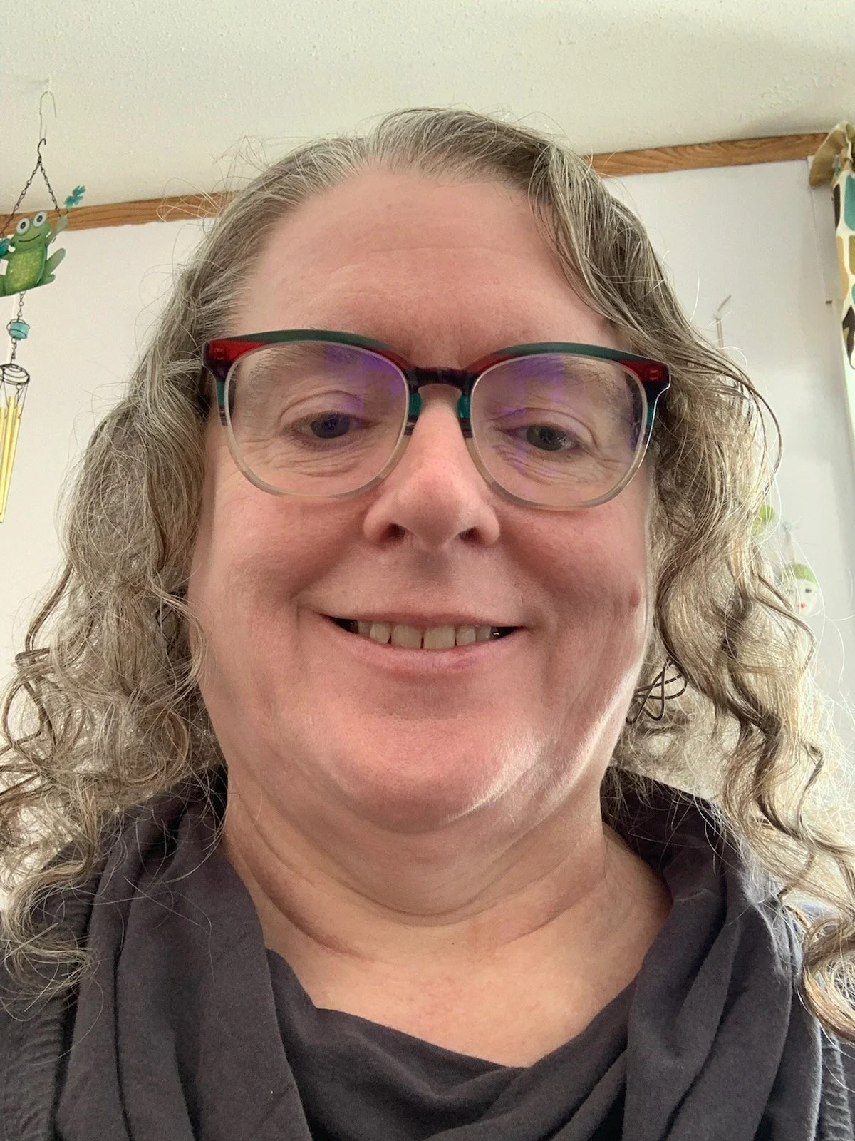 Close-up of a middle-aged woman, Trudy, with curly hair and glasses smiling indoors, with a frog-shaped hanging decoration visible in the background.