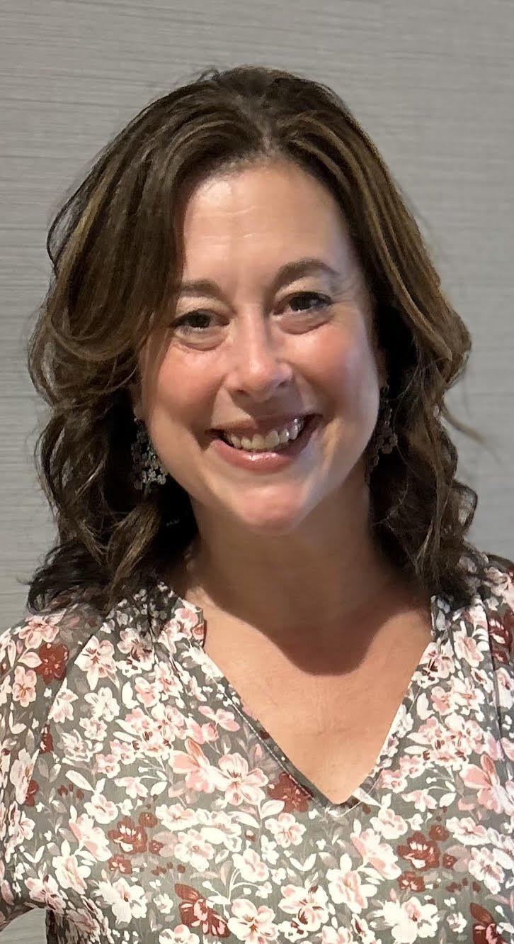 Professional photo of Bethany Hobbs, a woman with shoulder-length wavy brown hair, smiling broadly in a floral top against a neutral background.