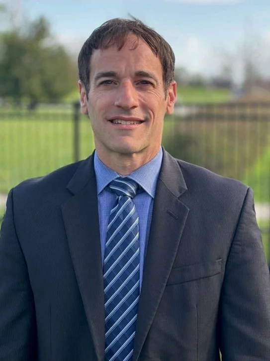 Professional photo of David Berman, a man with short dark hair, smiling warmly in a suit and striped blue tie, photographed outdoors against a green background.