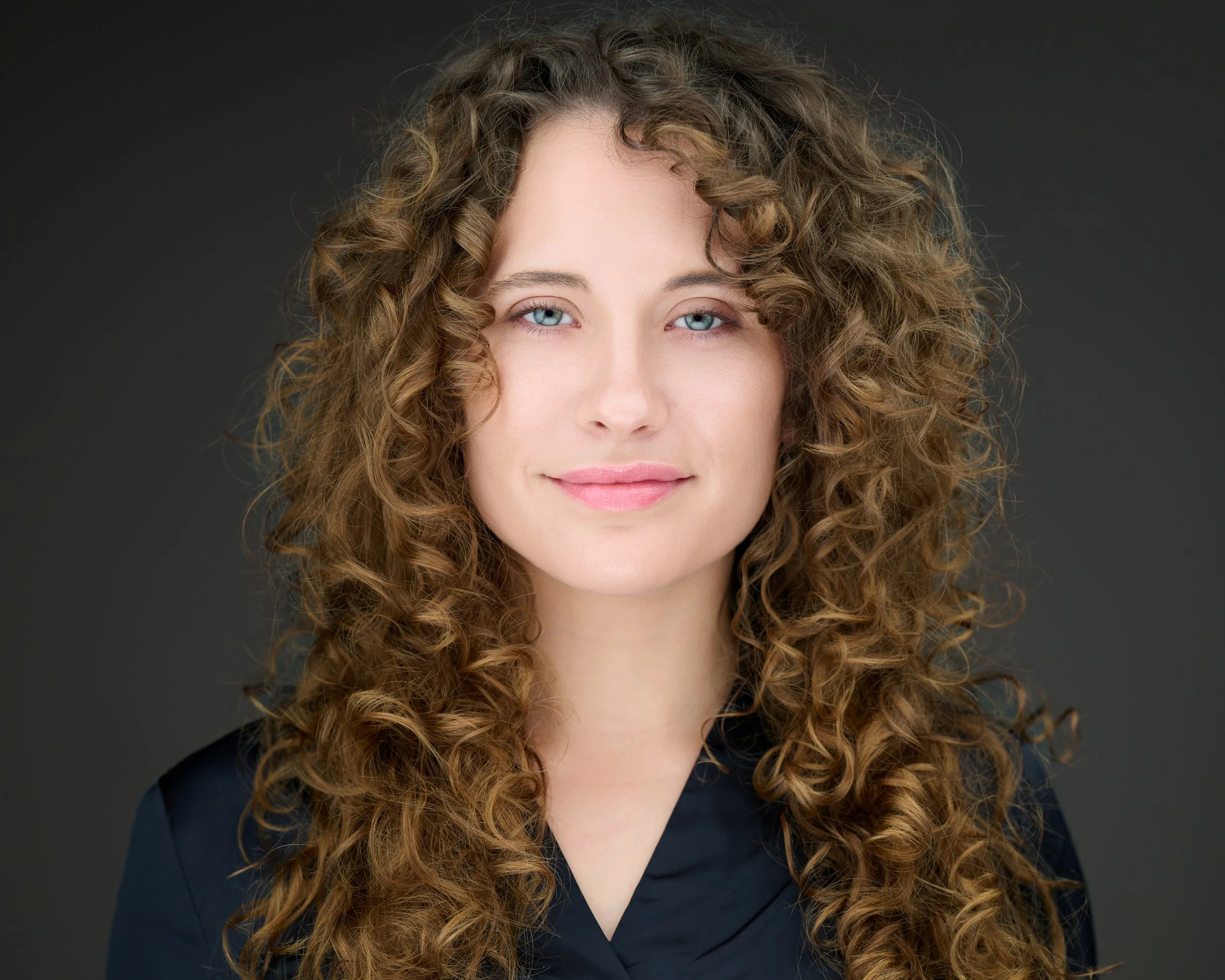 Professional headshot of Jess Trombly, a woman with long curly brown hair and blue eyes, smiling warmly against a neutral gray background.
