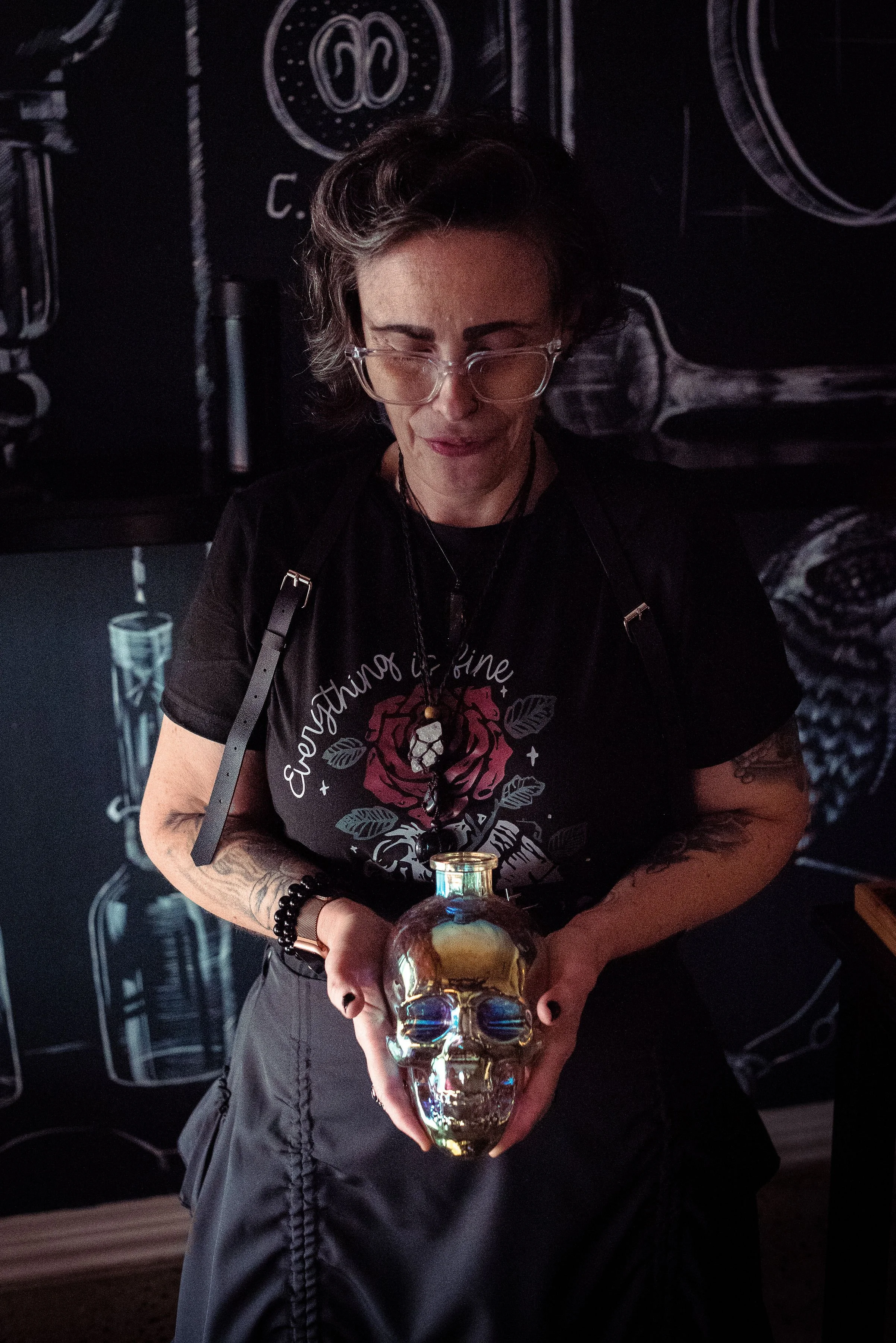 A woman wearing glasses and a black T-shirt with a rose design is holding a shiny skull-shaped object in front of her. The background has dark-colored artwork or chalk drawings of bottles and mechanical parts.