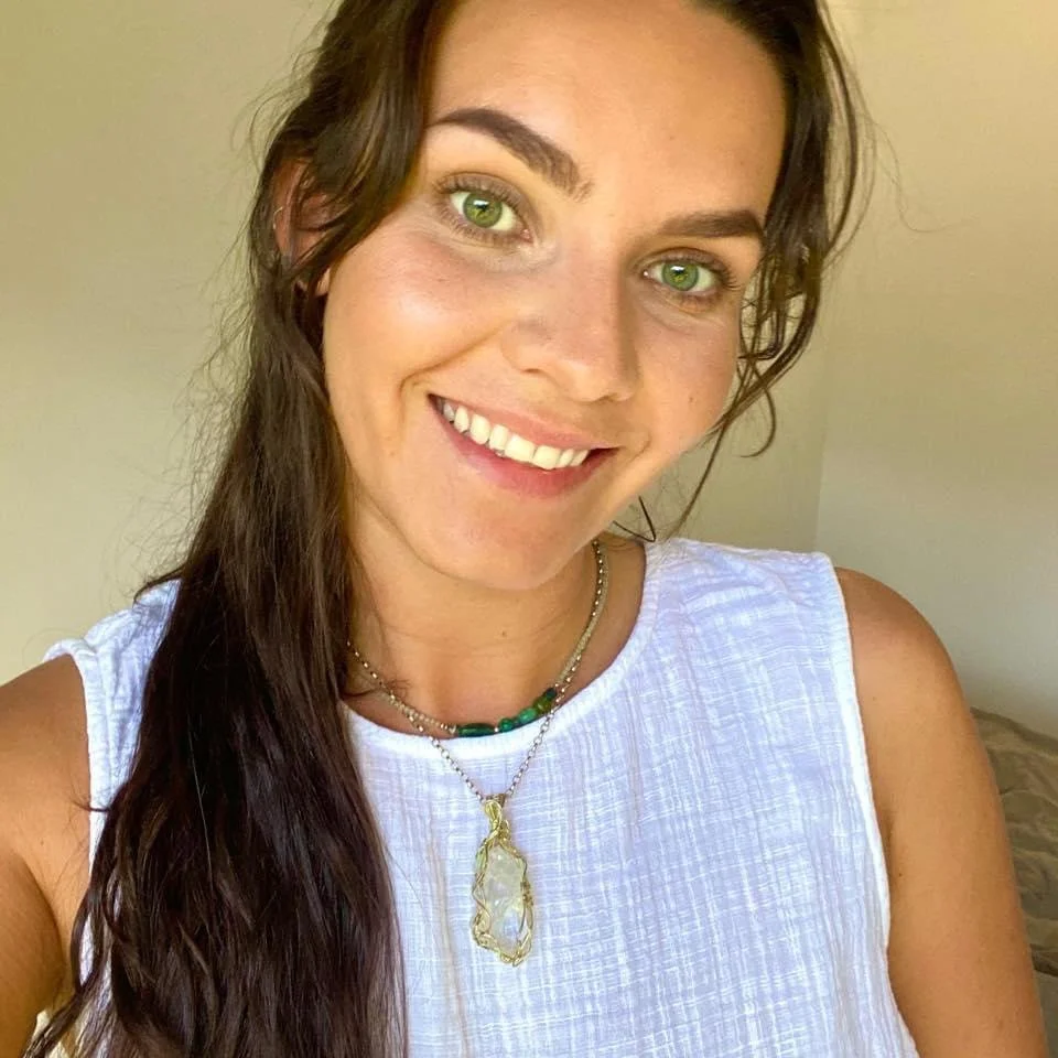 A young woman with long brown hair and green eyes smiling at the camera. She is wearing a white sleeveless top and two necklaces, one with a green bead and the other with an ornate pendant. The background is plain and neutral.