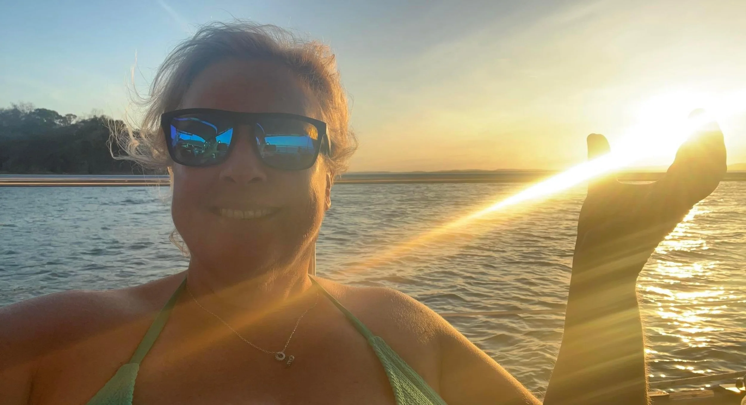 A woman smiling, wearing sunglasses, outdoors at sunset by the water, with sunlight reflecting off the water and lens flare in the scene.
