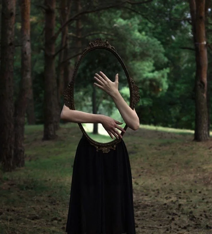 A person standing in a forest is holding an ornate mirror frame in front of their face, with their arms extended through the frame. The person's head is replaced by the mirror frame, creating a surreal appearance.