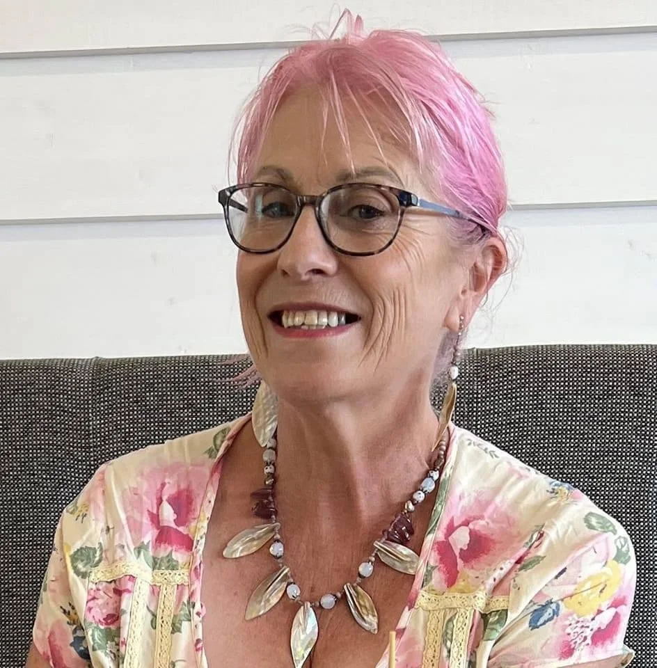A smiling woman with pink hair, glasses, and a floral dress, sitting on a textured gray couch, wearing a shell necklace and earrings in front of a white paneled wall.