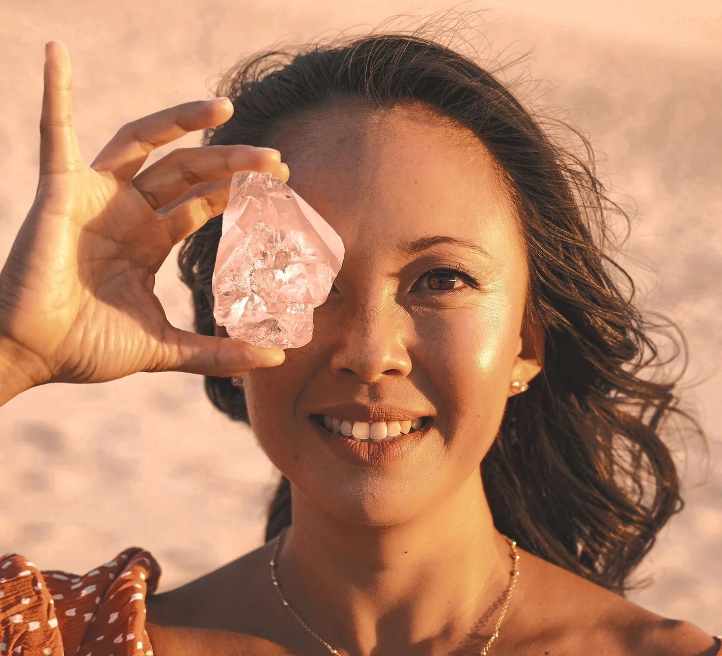A woman smiling at the camera holds a large pink crystal up to her right eye in a bright, outdoor setting.