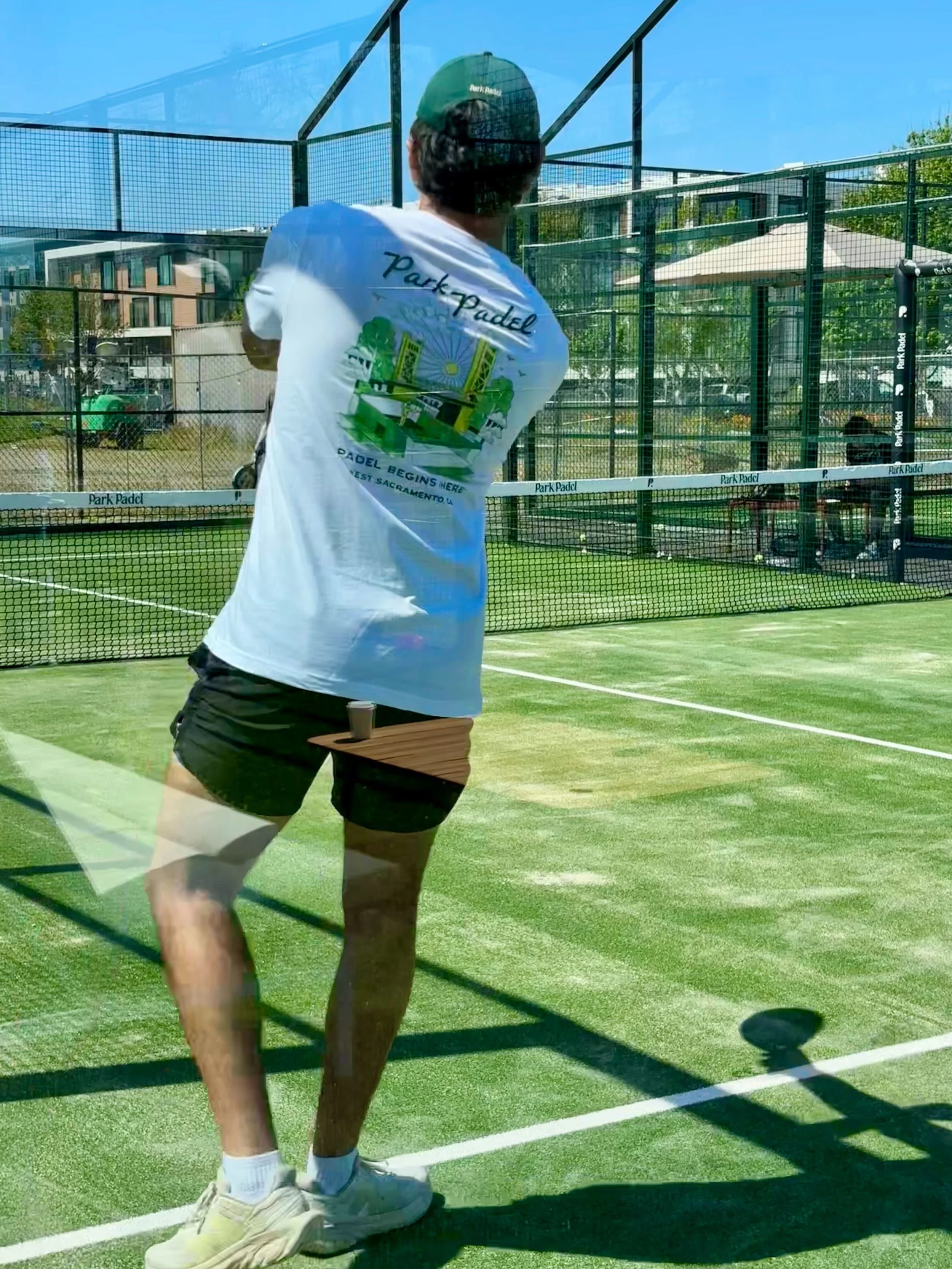 Person playing paddle tennis on an outdoor court, wearing a green cap, white T-shirt, black shorts, and white sneakers, with the court enclosed by a net and surrounded by buildings and trees.