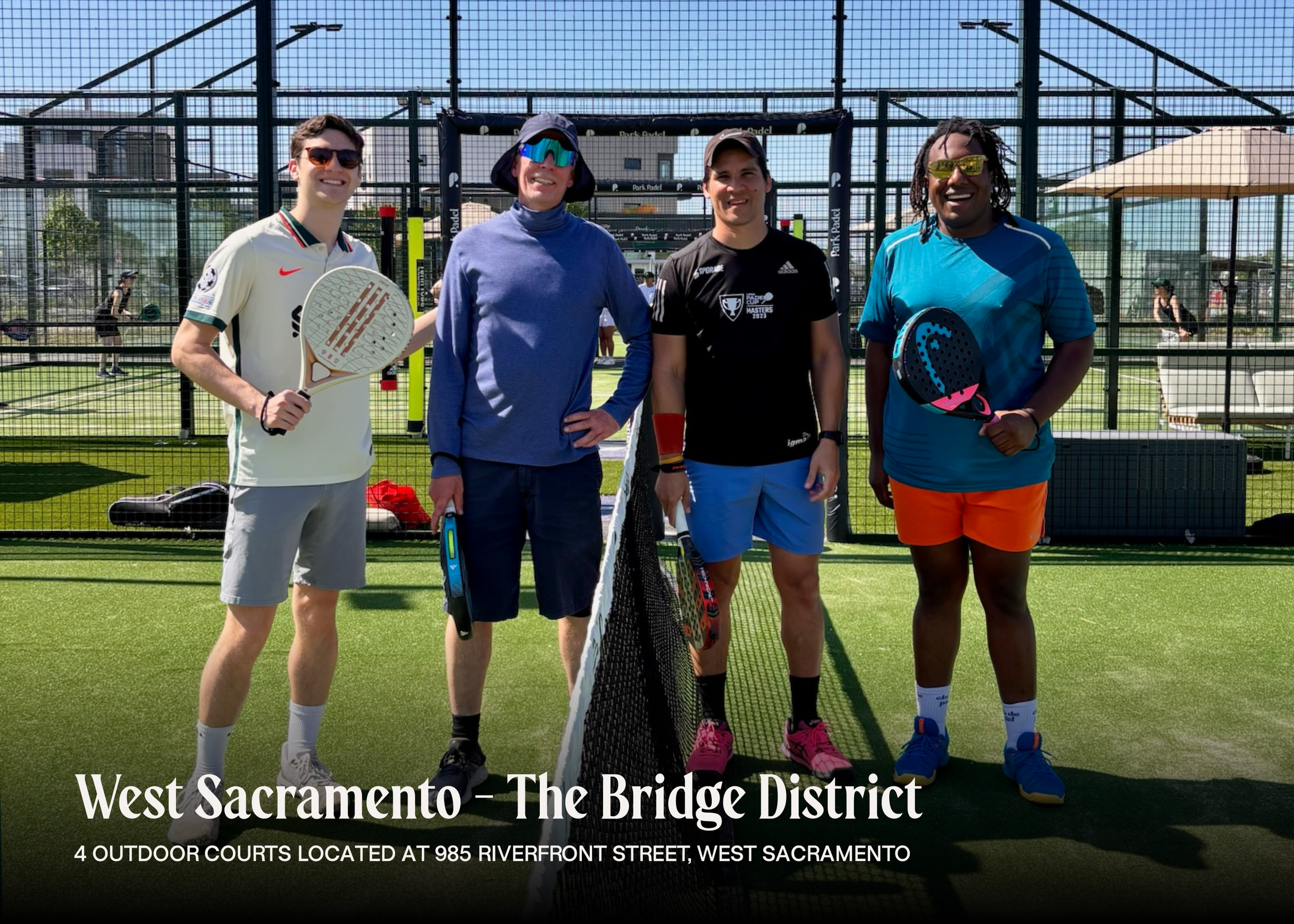 Four men standing on a padel tennis court, holding paddles, smiling, with a net in front of them. Two men are wearing sunglasses, one with a blue long sleeve, another with a black shirt, and the third man in a white shirt and gray shorts, the fourth 
