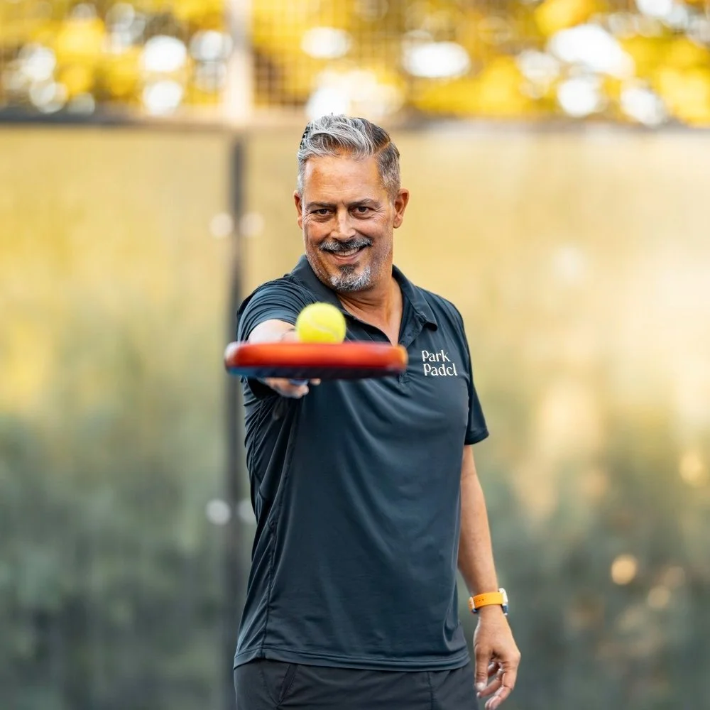 Smiling man in a grey shirt with 'Park Padel' logo, holding a red paddle with a yellow ball, standing outdoors with blurred yellow foliage background.