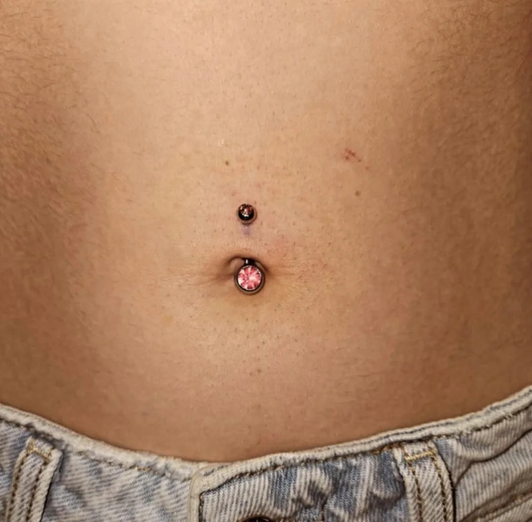 Close-up of a navel piercing with pink gemstones, above jeans waistband.