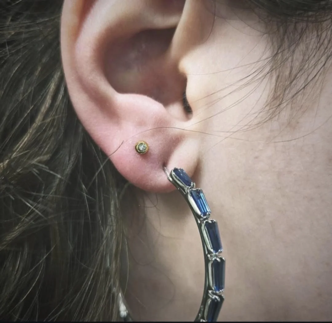 Close-up of a person's ear with two earrings. The first earring is a small stud, and the second is a large hoop with blue rectangular gemstones.