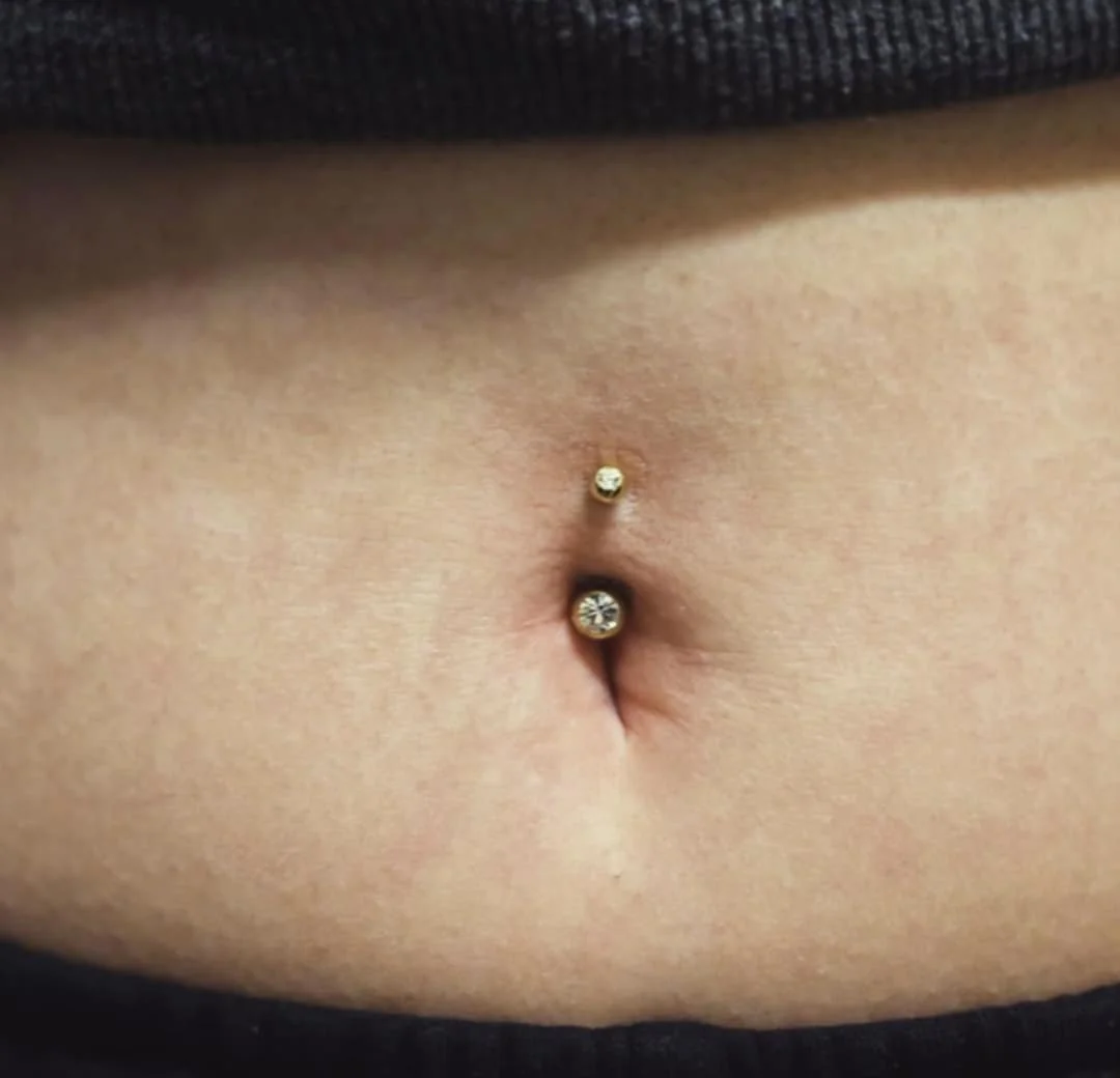 A close-up of a belly button with a gold navel piercing featuring two small gemstones.