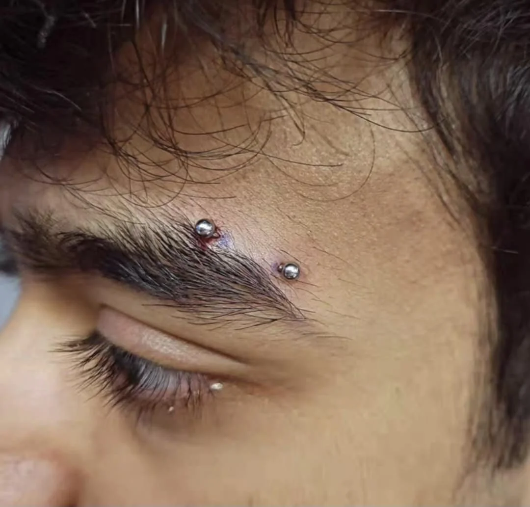 Close-up of a person's eyebrow with two metallic studs from a piercing and a smaller stud below the eye.