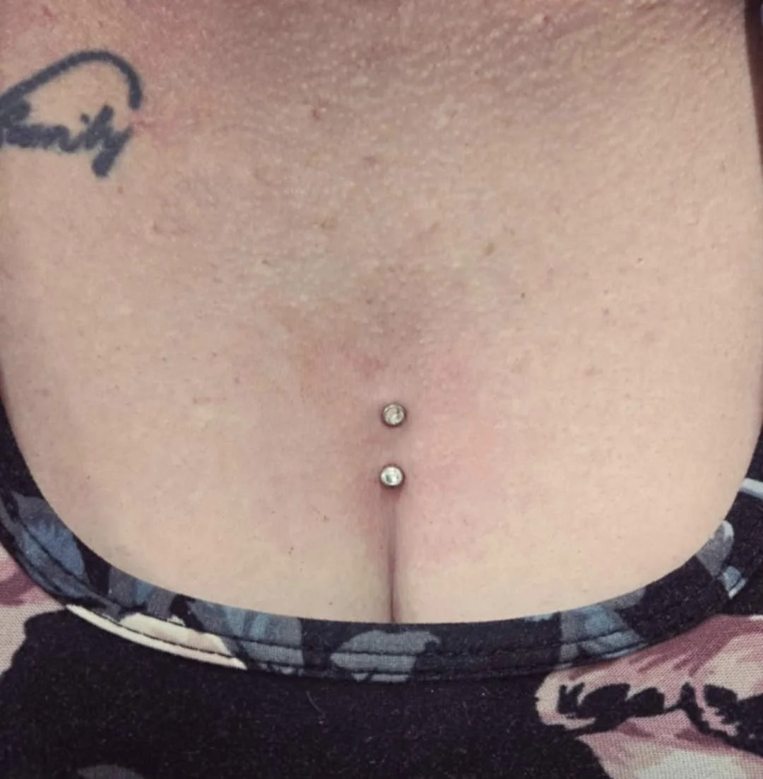 Close-up of a person's upper chest with two dermal piercings and a tattoo, wearing a floral-patterned garment.